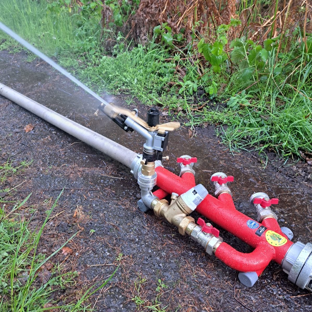 "Ein Feuerwehrmann bedient einen Waldbrandverteiler während eines Einsatzes im Wald. Der Verteiler ist an mehrere Schläuche angeschlossen, die Wasser zu verschiedenen Einsatzstellen leiten. Im Hintergrund sind Rauch und Bäume zu sehen, die auf die Bekämpfung eines Waldbrands hindeuten."
